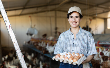 Latin female farmer in plaid shirt holding special tray of organic harvested chicken eggs and smiling at camera in henhouse