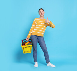 Smiling woman with shopping basket full of products showing thumbs up on light blue background
