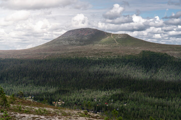 Städjan Mountain looms over a lush, dense boreal forest in Dalarna. Holiday houses nestle at its foothills in Idre, Sweden, bathed in summer sunlight under a cloudy sky