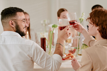 Young adult Caucasian woman and young adult Black man clinking glasses with other young adults during wedding celebration, smiling and engaging in toast at festive table