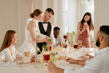 Caucasian bride and groom cutting wedding cake while diverse group of young adult guests sitting at table watching and smiling, young woman standing taking photo with smartphone