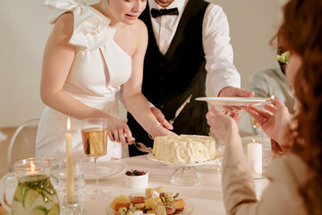 Caucasian young adult bride cutting wedding cake with help from Caucasian young adult groom while...
