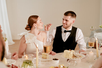 Caucasian young adult man feeding cake to Caucasian young adult woman during wedding celebration, both smiling and sitting at decorated table with desserts and drinks, other guests partially visible