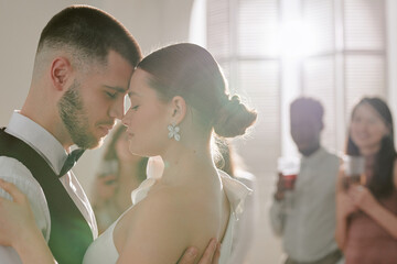 Caucasian young adult man and Caucasian young adult woman dancing closely during wedding celebration, eyes closed, multiethnic group of young adults watching in background