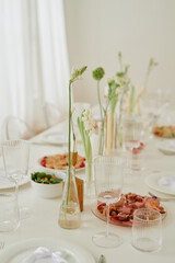 Elegant wedding celebration table setting featuring glassware, plates, floral arrangements and snacks arranged on tablecloth in bright indoor space