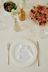 Closeup of elegant wedding table setting featuring plate with ribbon, cutlery, glassware, salad, charcuterie platter, floral decor, candle holder