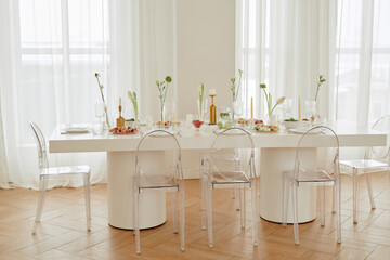 Empty wedding celebration table set with plates, glasses, candles, and floral arrangements standing in bright room, transparent chairs surrounding table, no people visible