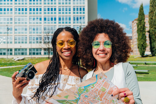 Female tourists holding city map and vintage camera, smiling while exploring urban landscape with modern architecture illuminating sunny day