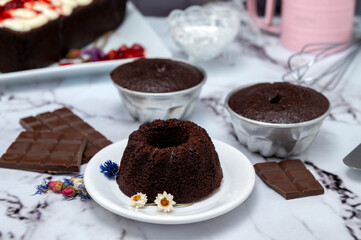 Rectangular and round chocolate pudding with cream on top and strawberries on a table with ingredients and pastry chef's hands decorating