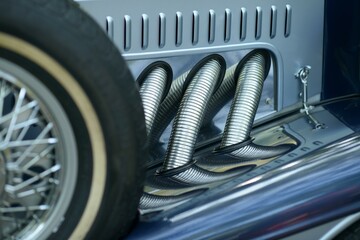 Side exhaust pipes and wire spare wheel with whitewall tire on a blue vintage luxury roadster polished chrome reflecting trees at an outdoor classic auto show