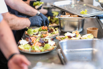 Close-up of chefs assembling fresh gourmet sandwiches with lettuce, cheese, and smoked salmon in a commercial kitchen. Hands wearing black gloves preparing food for catering service.