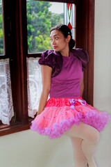 Asian young adult ballerina dressed up in colorful tutu skirt outfit sitting on window sill and looking afar through the window
