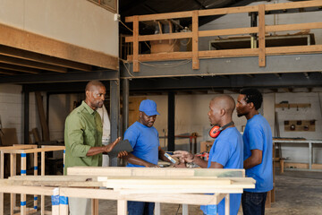 African American carpentry workers discussing measurements at shop workbench with tablet and tools