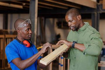 African American male coworkers measuring wood plank in carpentry shop with pencil goggles earmuffs