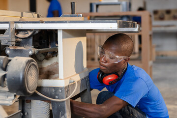 African American man kneeling in shop by table saw wearing goggles adjusting dust hose, copy space