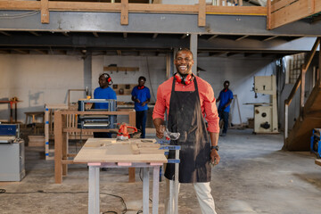 African American male coworkers working on wooden planks in workshop using power sander and clamps