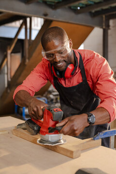 African American man using orbital sander on bench in wood shop wearing goggles and ear protection