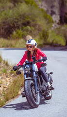 Young motorcyclist wearing a red jacket and tights taking a curve on his customized chopper-type motorcycle along a mountain road on a sunny day.