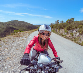 Front view from the handlebars of a smiling female motorcyclist riding a customized chopper along a mountain road on a sunny day.