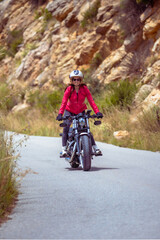 A smiling young motorcyclist in a red jacket riding standing on his customized chopper motorcycle along a rocky mountain road on a sunny day.