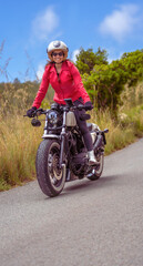 A young, smiling, happy female motorcyclist wearing a red jacket and open-face helmet riding her customized chopper motorcycle on a steep mountain road, standing on the footrests under a blue sky.