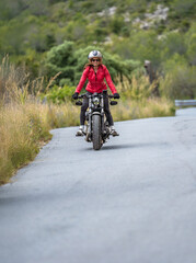 A young, smiling, happy female motorcyclist wearing a red jacket, tights, and an open-face helmet riding her customized chopper motorcycle down a steep mountain road, standing on the footrests.