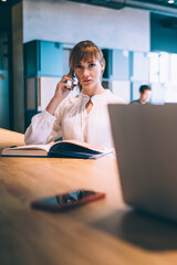 Adult determined businesswoman making phone call near pleased coworker working on laptop in office