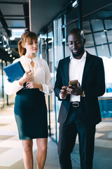 Pensive diverse coworkers interacting with smartphone in corridor of cozy modern office