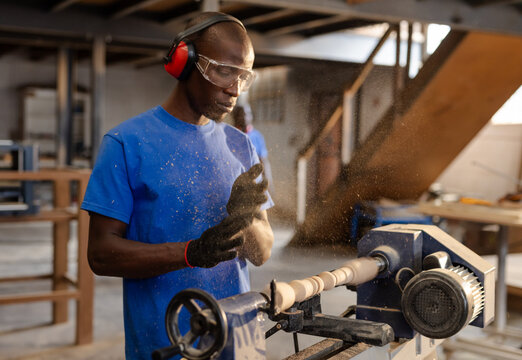 African American man operating lathe wearing goggles at carpentry workshop shaping wooden spindle