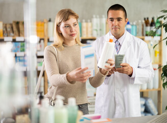 Careful young woman holding cream and man apothecary consulting her in drugstore