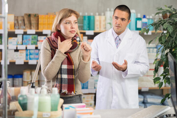 Young woman with a cold throat and a wrapped scarf chooses cough medicines and antipyretics against the background of a pharmacist in a pharmacy. Buying antiviral medications