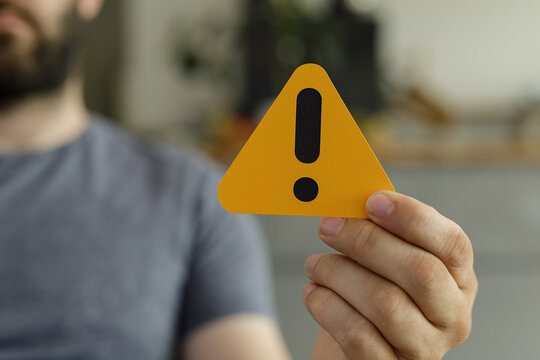 Close-up of a man's hand holding a yellow triangle with an exclamation mark, signifying caution or warning.
