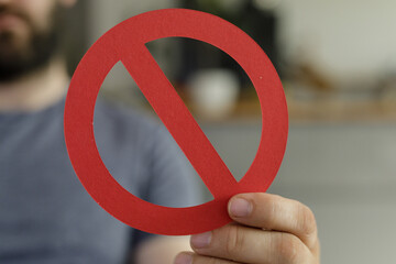 Close-up of a hand holding a red circle with a diagonal line through it, symbolizing prohibition or...