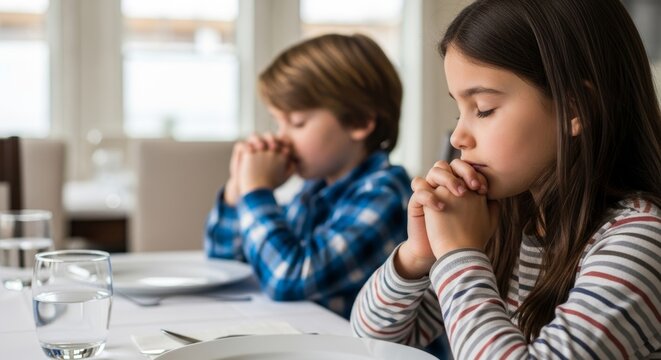 Medium close-up of two children sitting at a dining table with a white tablecloth, holding hands and praying before their meal. A heartfelt moment of gratitude and family tradition.