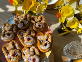 Bunny shaped cinnamon buns, with a dried cranberry as the nose, topped with icing and pastel sprinkles. These Easter treats sit on a breakfast table, next to springtime daffodil flower arrangements. 