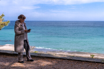 60 year old woman in quilted coat against beautiful seascape with turquoise water. Woman checks messages on mail on phone. Calmness and solitude for travel blog.
