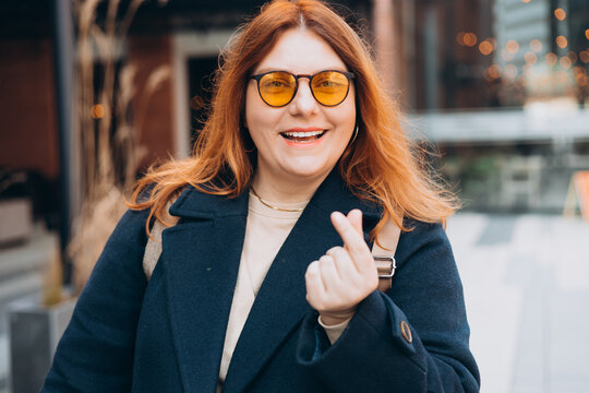 Female hand showing finger heart or mini heart gesture with the index finger and thumb come together like a snap outdoors. 30s Woman in sunglasses on urban background. High quality photo.