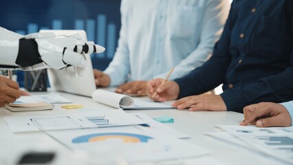 A diverse business team engaged in collaborative work with the assistance of a robotic arm, analyzing data and strategizing in a modern office setup.Noogenesis