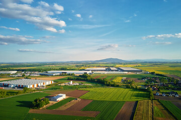 Aerial view of industrial warehouse complex near green agricultural fields and small village. Modern logistics infrastructure in rural landscape