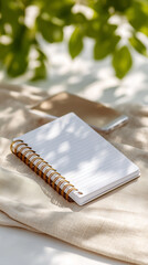 Blank notebook and smart phone on table is bathed in sunbeam shining through lush green leaves, evokes feeling relaxation and creative inspiration.