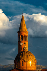 A bell tower and dome of a Moretta church in the warm light of a sunset, set against a dramatic sky with white clouds in the background in Alba, Italy.