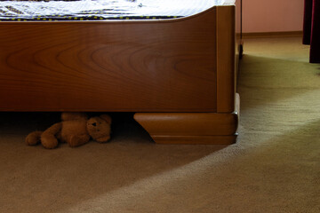 Teddy bear lies on the floor under a wooden bed in the bedroom, a child's toy on a carpet, home...