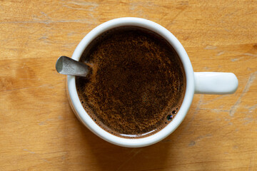 A cup of black coffee with foam and a spoon in a white mug on a wooden table, top view, a morning drink for energy