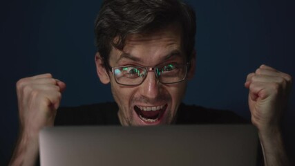 Excited crypto trader wearing glasses with bitcoin charts reflected in the lenses celebrating cryptocurrency market success, reacting with excitement, raising his fists looking at a laptop screen. 