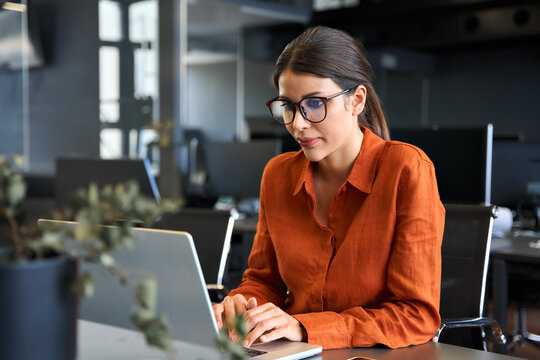 European mature woman in eyeglasses using computer for finance data analysing work online. Focused financial specialist latin hispanic businesswoman working on laptop pc sitting at desk in office. 