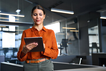 Latin Hispanic mature adult professional business woman looking at online trade app. European businesswoman CEO holding digital tablet using fintech tab application standing at workplace in office.