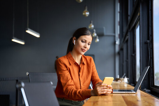 Young Indian business woman manager ceo using cell phone mobile app laptop. Smiling Latin Hispanic mature female businesswoman holding smartphone in office trading, reading, working online. Copy space - Powered by Adobe