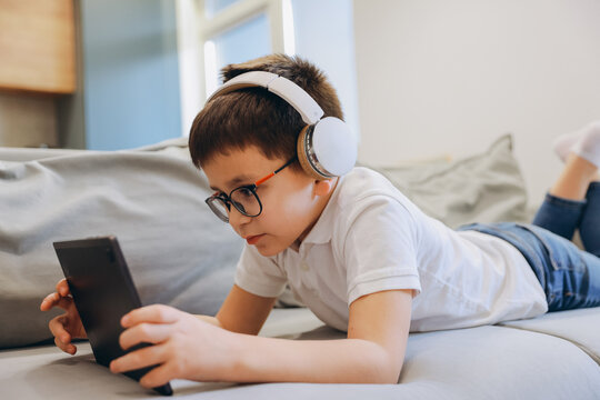Young boy with headphones and eyeglasses using tablet while lying on sofa at home