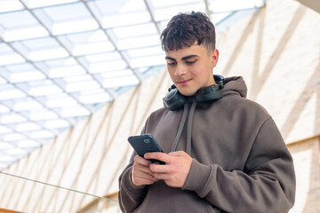 Young man using smartphone with headphones around neck in modern glass building, teenager texting...
