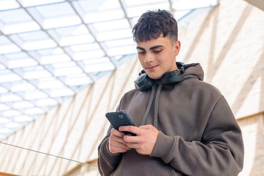 Young man using smartphone with headphones around neck in modern glass building, teenager texting on mobile phone in contemporary architecture setting with natural light. Copyspace.  - Powered by Adobe